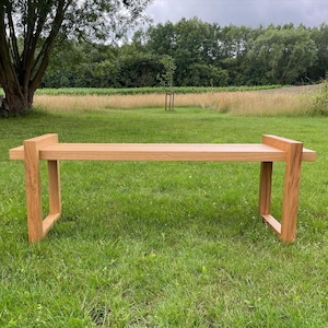 May include: A light brown wooden bench with a rectangular seat and geometric legs. The bench is outdoors, set on a green grassy lawn. The bench is approximately 60 inches long.