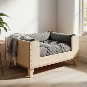 K&ouml;nnte beinhalten: Ein helles Hundebett aus Holz mit einem grauen Kissen und Kissen. Eine graue Decke mit Fransen ist &uuml;ber die Seite drapiert. Das Bett hat ein modernes Design mit sichtbaren Verbindungen und abgewinkelten Beinen. Das Bett befindet sich in einem Raum mit einem Fenster und einer Pflanze.