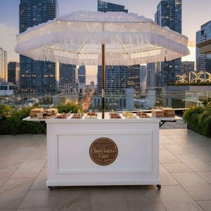 Puede incluir: Un carrito de charcutería blanco con una gran sombrilla con flecos, sobre un fondo de paisaje urbano. El carrito presenta un letrero de madera que dice "Charcuterie Cart". Tiene múltiples compartimentos llenos de alimentos y dos mesas laterales.