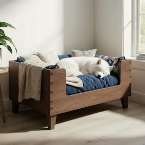 K&ouml;nnte beinhalten: Ein Hundebett aus Holz mit dunkler Walnuss-Oberfl&auml;che. Das Bett hat ein blaues Kissen, dekorative Kissen und eine beige Decke. Ein wei&szlig;er Hund schl&auml;ft auf dem Bett. Das Bett befindet sich in einem hellen Raum.
