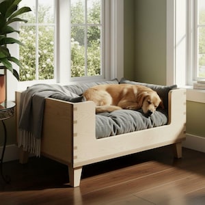 K&ouml;nnte beinhalten: Ein helles Hundebett aus Holz mit einem grauen Kissen und einer grauen Decke. Ein Golden Retriever liegt auf dem Bett. Das Bett steht in der N&auml;he eines Fensters mit Blick auf B&auml;ume.