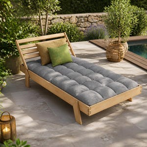 May include: A wooden outdoor chaise lounge with a gray tufted cushion and two decorative pillows. The lounge has a slatted back and is set on a stone patio. A wicker basket and a lit lantern are in the background.