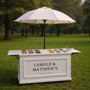 Pu&ograve; includere: Un carrello alimentare bianco con un ombrellone bianco e beige. Il carrello ha un piano di lavoro con scomparti per alimenti e un cartello con la scritta "CAMILLE & MATTHEW'S Wedding Weekend". Il carrello &egrave; su ruote ed &egrave; su un prato.
