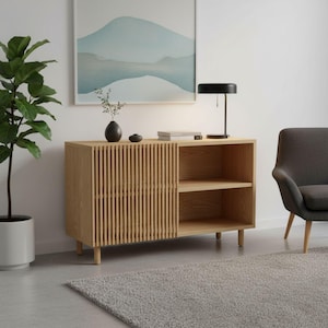 Könnte beinhalten: Ein helles Holzschrank mit einer Lamellentür und offenen Regalen. Eine schwarze Lampe, eine kleine Vase und dekorative Gegenstände stehen obenauf. Ein grauer Sessel und eine Topfpflanze befinden sich ebenfalls im Raum. Ein hellgrauer Teppich bedeckt den Boden.