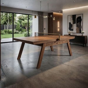 Könnte beinhalten: Ein moderner Tischtennistisch aus Holz mit Netz in einem hellen Raum. Der Tisch hat eine dunkle Holzoberfläche und schräge Beine. Eine Schublade mit Messingknopf ist sichtbar. Der Raum verfügt über große Fenster, moderne Kunst und Pendelleuchten. Der Tisch ist ca. 274 cm lang.