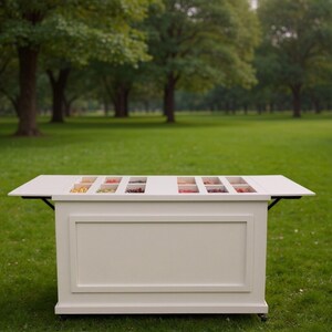 Pu&ograve; includere: Un buffet bianco con pi&ugrave; scomparti per alimenti, posizionato all'aperto su un prato. Il tavolo ha un design rettangolare con un pannello inferiore e un piano pieghevole. Lo sfondo presenta alberi.