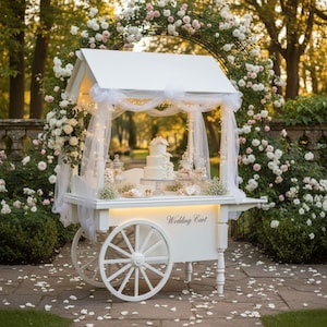 Puede incluir: Un carrito de boda blanco con techo a dos aguas, adornado con tela transparente y luces de cadena. El carrito exhibe una tarta de varios pisos, caramelos y pasteles. Las palabras "Wedding Cart" están impresas en la parte delantera. El carrito está rodeado de flores.