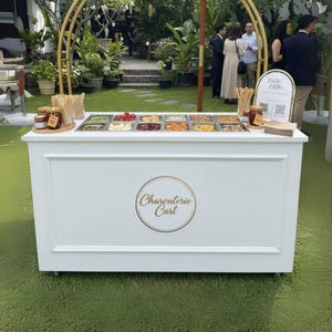 May include: A white charcuterie cart with a gold logo reading "Charcuterie Cart". The cart features a top section with various food items in stainless steel containers. The cart is on wheels and is set up outdoors on grass.