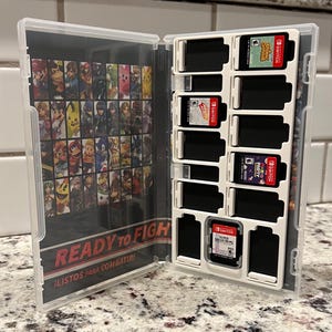 May include: A clear plastic case with a black and white image of video game characters on the front cover. The case has 12 slots for game cartridges, and the text "READY TO FIGHT" and "LISTOS PARA COMBATIR" is printed on the front cover.