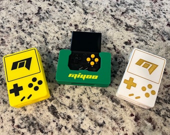 May include: Three handheld game consoles in different colors. The console on the left is yellow with black buttons and a black "M" logo. The console in the middle is green with a black screen and yellow buttons. The console on the right is white with gold buttons and a gold "M" logo. The console in the middle has the word "Miyoo" printed on it.