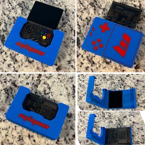 Op de afbeelding: Een blauwe en rode 3D-geprinte case voor een draagbaar spelapparaat. De case heeft een uitsparing voor het scherm en de knoppen van het apparaat, en het woord "Miyoo" is in rode letters op de voorkant gedrukt.
