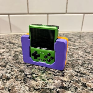 May include: A green handheld video game console with a purple and orange stand. The console has a screen, buttons, and the text "GAME KIDDY  HND-101" on the front.
