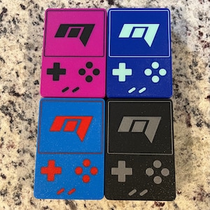 Op de afbeelding: Vier kleine draagbare gameconsoles met verschillende gekleurde behuizingen. De consoles zijn blauw, zwart, roze en blauw. Elke console heeft een ander gekleurd logo en knoppen. De consoles zijn allemaal in dezelfde stijl en hebben een vergelijkbaar ontwerp.