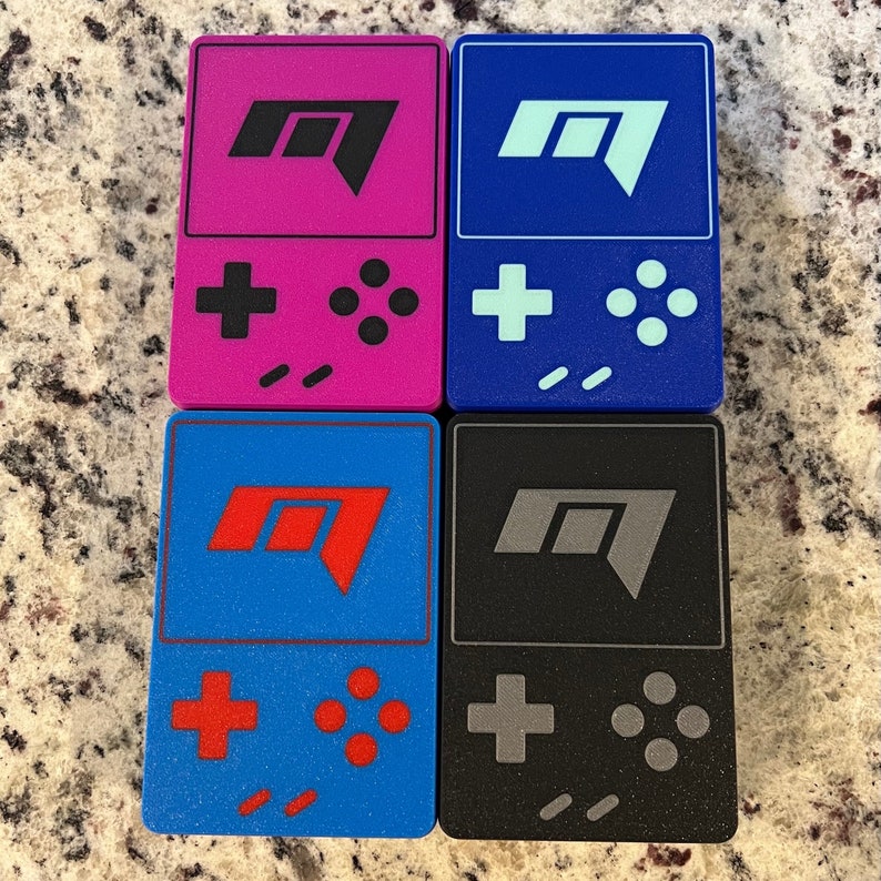 Op de afbeelding: Vier kleine draagbare gameconsoles met verschillende gekleurde behuizingen. De consoles zijn blauw, zwart, roze en blauw. Elke console heeft een ander gekleurd logo en knoppen. De consoles zijn allemaal in dezelfde stijl en hebben een vergelijkbaar ontwerp.