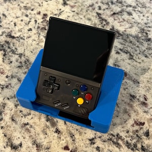 Op de afbeelding: Een zwarte handheld-gameconsole met een blauwe standaard. De console heeft een scherm, een directionele pad, vier gekleurde knoppen en knoppen met de labels "Select", "Start", "A", "B" en "Y".