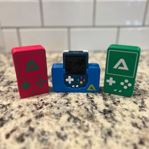 Op de afbeelding: Drie draagbare gameconsoles, een rode, een blauwe en een groene, elk met een gestileerde letter 'A' en een directionele pad en knoppen.