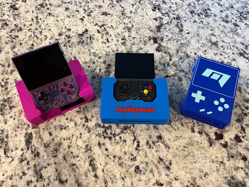 Op de afbeelding: Drie draagbare gameconsoles in beschermende hoesjes. De eerste console is paars en heeft een zwart scherm. De tweede console is zwart en heeft een blauwe hoes met het woord "analogue" erop gedrukt. De derde console is blauw en heeft een wit "M"-logo erop.