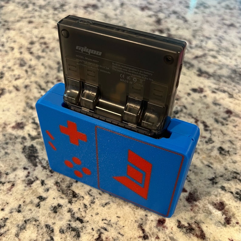 Op de afbeelding: Een blauwe en rode handheld-gameconsole met een zwarte cartridge erin. De console heeft een plusteken, vier stippen en een rode letter D aan de voorkant.
