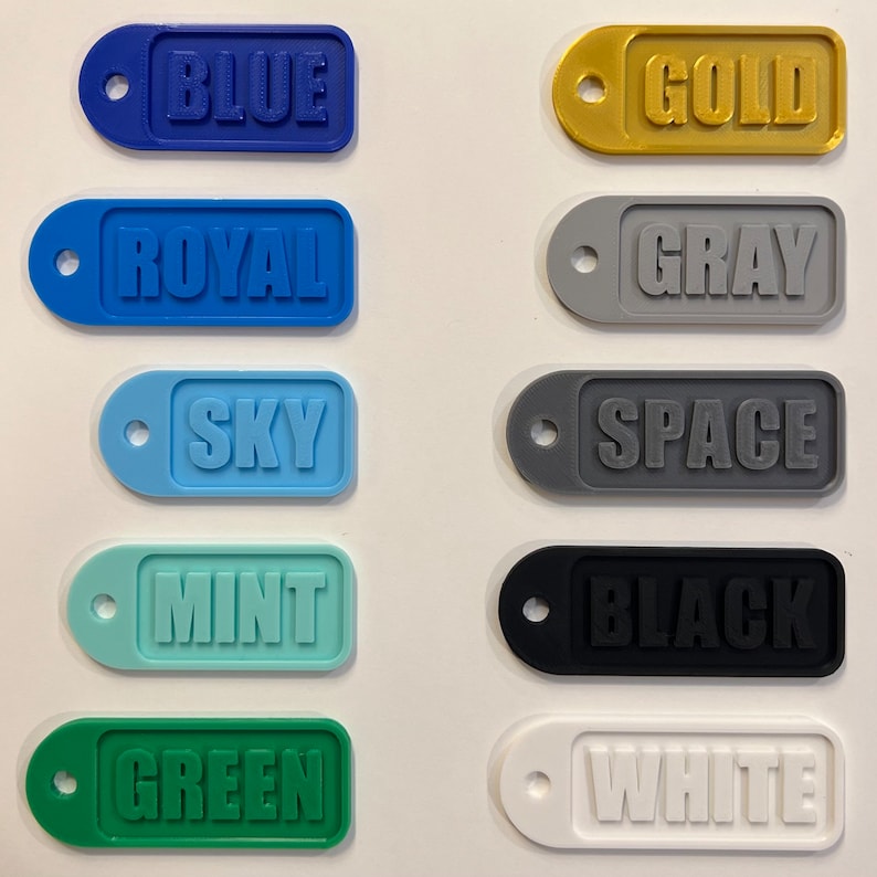 Op de afbeelding: Tien plastic sleutelhangers in verschillende kleuren met tekst. De kleuren zijn blauw, koninklijkblauw, hemelsblauw, mintgroen, groen, goud, grijs, spacegrijs, zwart en wit. De tekst op de sleutelhangers is "BLUE", "ROYAL", "SKY", "MINT", "GREEN", "GOLD", "GRAY", "SPACE", "BLACK" en "WHITE".