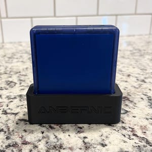 May include: A blue handheld gaming device with a black stand. The stand has the word "ANBERNIC" printed on the front.