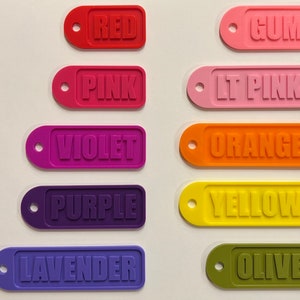 Op de afbeelding: Tien kleurrijke rechthoekige labels met afgeronde hoeken en een gat bovenaan. De labels zijn gelabeld met de volgende kleuren: Rood, Roze, Violet, Paars, Lavendel, Gom, Lichtroze, Oranje, Geel, Olijf.