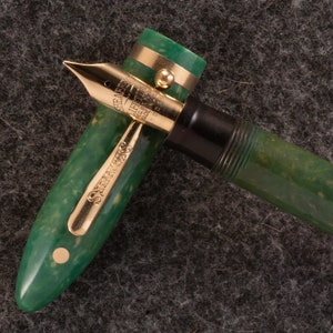 May include: A green and gold fountain pen with a gold nib and a black cap. The pen is on a gray surface.