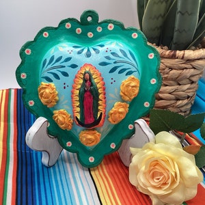 May include: A green heart-shaped ceramic ornament with a blue and white painted design featuring a religious figure and floral accents. The ornament is on a white stand and a colorful striped surface.