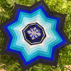 May include: A handmade, star-shaped wall hanging with a layered design. The piece features a gradient of blues, from dark navy to light turquoise, with a black and white central design. The artwork is displayed against a backdrop of green foliage.