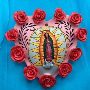 May include: A pink heart-shaped ceramic tile with a painted image of the Virgin Mary in a blue and red robe. The tile is surrounded by 10 red roses.