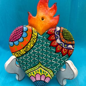 May include: A vibrantly painted wooden heart with a flame design on top. The heart is teal with colorful floral patterns and rests on a white stand. The flame is orange and red.