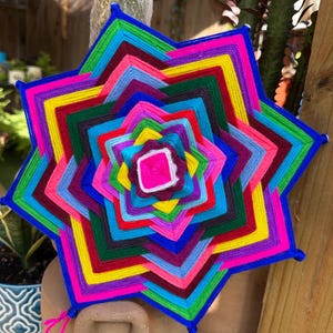 May include: A vibrant, multi-colored star-shaped yarn art piece. The artwork features concentric layers of bright colors including pink, yellow, green, blue, and purple. The edges are outlined in blue, and the center has a white and pink square.