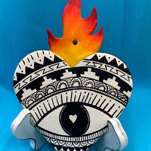 May include: A heart-shaped decorative piece with a flame on top. The heart is white with black geometric patterns and an eye design. It is supported by a small, white stand. The flame is painted in shades of red, orange, and yellow.
