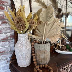 May include: A white ceramic vase with a textured pattern holds dried yellow flowers. A brown wooden bead garland hangs from the vase and a brown ceramic vase with a textured pattern sits on a dark wood surface.