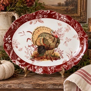 May include: An oval ceramic serving platter with a scalloped edge featuring a deep red border decorated with white botanical illustrations. The center of the platter displays a detailed, colorful illustration of a turkey standing in grass.