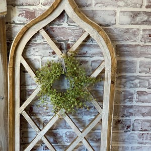 Puede incluir: Un marco de ventana decorativo de madera con un dise&ntilde;o de celos&iacute;a de diamantes. El marco est&aacute; pintado de blanco con bordes desgastados, y una peque&ntilde;a corona verde cuelga en el centro. El fondo es una pared de ladrillo.
