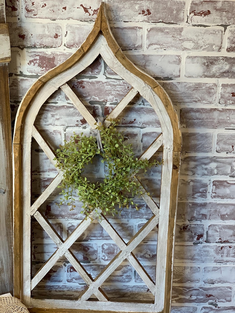 Puede incluir: Un marco de ventana decorativo de madera con un dise&ntilde;o de celos&iacute;a de diamantes. El marco est&aacute; pintado de blanco con bordes desgastados, y una peque&ntilde;a corona verde cuelga en el centro. El fondo es una pared de ladrillo.