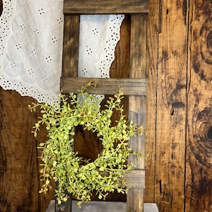 May include: A rustic wooden ladder shelf with a green leafy wreath hanging on the middle rung. The ladder is stained dark brown and has a distressed look. The wreath is made of artificial greenery and has a natural, organic feel.