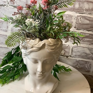 May include: White ceramic planter shaped like a woman's bust with a crown of leaves and pink and white flowers.