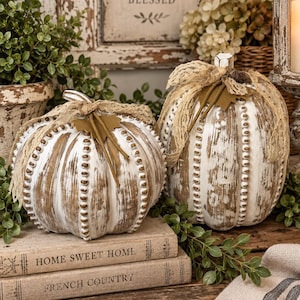 May include: Two decorative pumpkins with a distressed white and brown finish, featuring vertical beaded ridges. Each pumpkin is topped with a tan lace ribbon and a small brown felt leaf. They are positioned on two stacked books labeled Home Sweet Home.