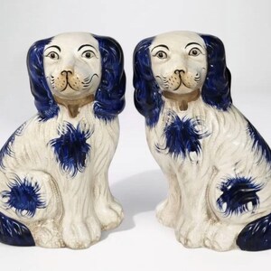 May include: Pair of ceramic dog figurines. Each dog is seated, with a white body and blue accents on the ears, tail, and chest. They have brown collars and detailed facial features, creating a classic decorative item.