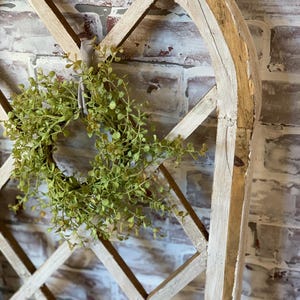 Puede incluir: Un arco decorativo de madera con un dise&ntilde;o de celos&iacute;a de diamantes, pintado con un acabado blanco envejecido. Una peque&ntilde;a corona de hojas verdes est&aacute; suspendida del arco con una cinta de color neutro. El fondo es una pared de ladrillo.
