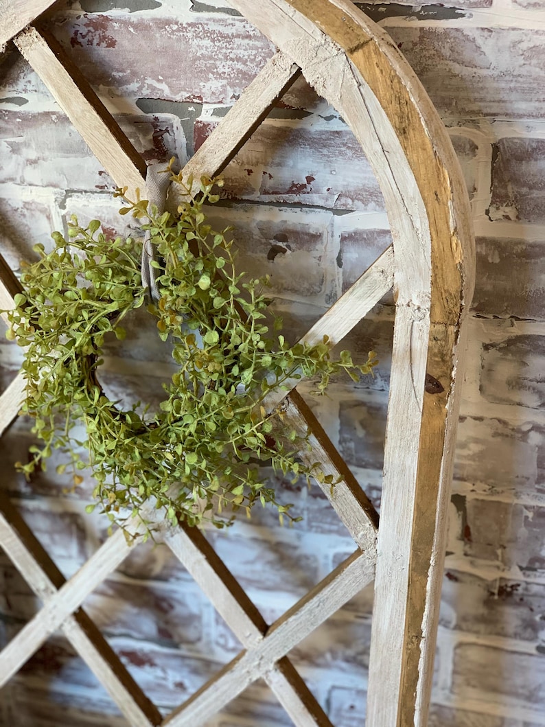 Puede incluir: Un arco decorativo de madera con un dise&ntilde;o de celos&iacute;a de diamantes, pintado con un acabado blanco envejecido. Una peque&ntilde;a corona de hojas verdes est&aacute; suspendida del arco con una cinta de color neutro. El fondo es una pared de ladrillo.