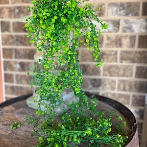 May include: A long strand of artificial greenery with small, round, green leaves. The greenery is hanging from a decorative stand and cascading down onto a wooden barrel.