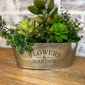 May include: A silver metal planter with the text "FLOWERS & GARDEN" on the front. The planter is filled with a variety of green succulents and plants.