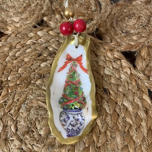 May include: Hand-painted Christmas ornament made from an oyster shell. The shell is edged in gold paint and features a Christmas tree design with a red bow. The ornament is suspended by a gold string and red and gold beads.