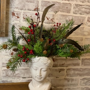 May include: A white ceramic bust vase filled with evergreen branches, red berries, and dark feathers. The bust has a serene expression and is placed on a wooden surface. The background is a white brick wall.