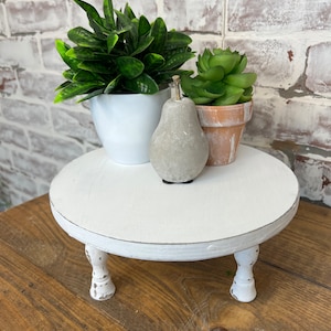 May include: White wooden tiered stand with three legs. The stand is decorated with a distressed white paint finish. The stand is topped with two potted plants, one with green leaves and the other with green succulents, and a gray concrete pear.