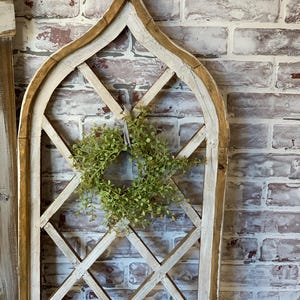 Puede incluir: Un marco de ventana de madera arqueado y envejecido con un dise&ntilde;o de celos&iacute;a de diamantes. El marco est&aacute; pintado de blanco con detalles en marr&oacute;n y presenta una peque&ntilde;a corona verde en el centro. El fondo es una pared de ladrillo.