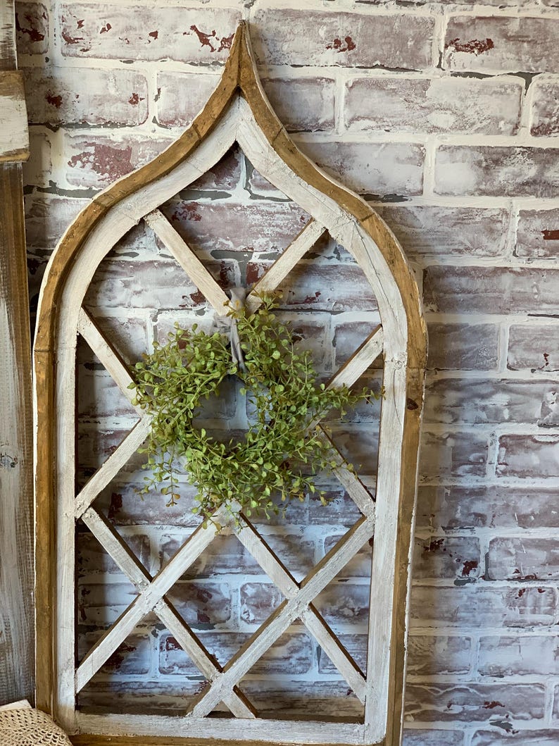 Puede incluir: Un marco de ventana de madera arqueado y envejecido con un dise&ntilde;o de celos&iacute;a de diamantes. El marco est&aacute; pintado de blanco con detalles en marr&oacute;n y presenta una peque&ntilde;a corona verde en el centro. El fondo es una pared de ladrillo.