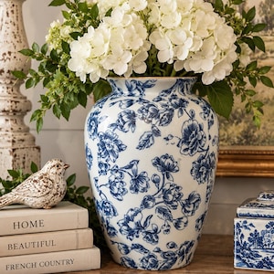 May include: A white ceramic vase featuring a blue floral pattern, filled with a large bouquet of white hydrangea flowers and green leaves. The vase is positioned next to a stack of three books labeled HOME, BEAUTIFUL, and FRENCH COUNTRY.